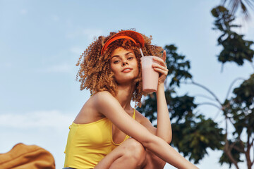 Young woman with curly hair enjoys a smoothie outdoors under a bright sky