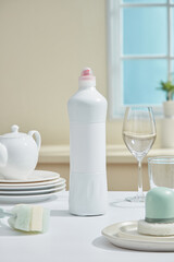 Close-up of a white bottle and ceramic plates neatly arranged on a beige surface, surrounded by soft window light and pastel tones. Kitchen dishwashing and cleaning products