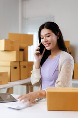 Asian woman entrepreneur managing retail business from home office talking on phone and calculating costs