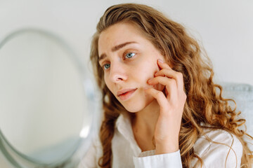 A cute woman examines her rashes in a small mirror in a bright room. A beautiful woman enjoys her...