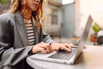 Portrait of a woman in stylish clothes at a table on a terrace working on a laptop. Smiling woman freelancer enjoying coffee in a coffee shop with a work computer at the table. Business concept.
