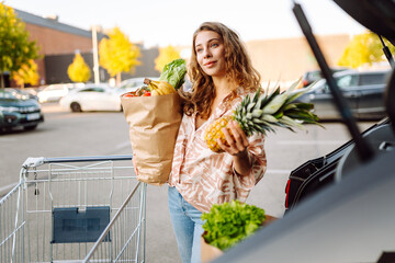 Young woman with shopping cart full of vegetables and fruits standing near car in parking lot. Housewife woman loading groceries into cars after grocery shopping. Consumerism concept.