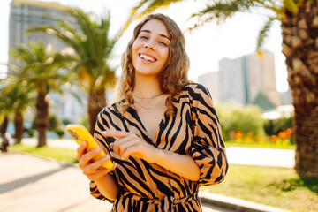 Happy woman enjoying sunny day in city park with phone. Young tourist with phone on walk, chatting...