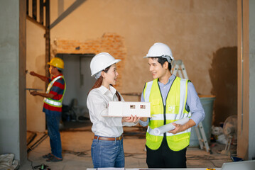 Asian engineers at building site reviewing architecture model. Wearing hard hats and safety vests, they discuss layout, planning, teamwork