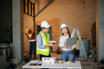 Asian engineers discuss floor plan using laptop and building model at active construction site. Collaboration, safety