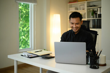 Confident businessman smiling while working on a laptop in a modern office with natural light