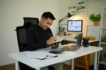 A Mature Asian businessman smiles while taking notes in a notebook at a modern office desk, with a laptop and documents around