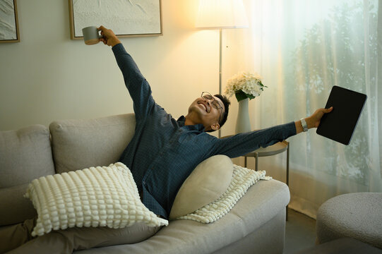 A joyful mature man stretches on a sofa, holding a tablet and a coffee mug, celebrating success or enjoying a moment of relaxation in his cozy modern living room - Powered by Adobe