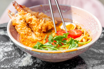 A bowl of ramen with tempura shrimp, cherry tomatoes, cilantro, and a soft-boiled egg, presented in bright daylight. The dish is vibrant and appetizing, highlighting diverse textures and colors