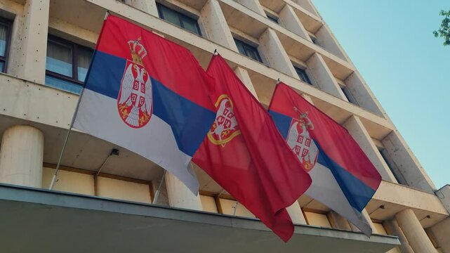 Serbian flags sway gently in a soft spring breeze, mounted on a Yugoslav-style government building under a clear blue sky
