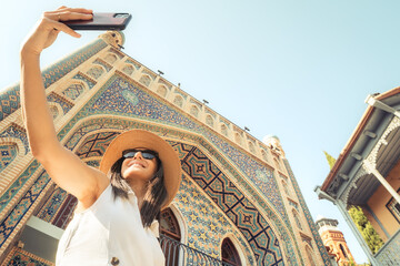 Woman taking selfie in front colorful Persian-style mosque building in Abanotubani district on summer day, smiling under sun, enjoying moment, concept freedom travel joy confidence