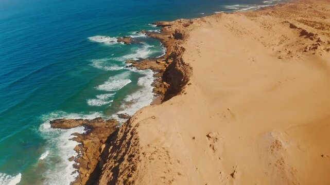 Aerial drone shot over the Tamri coastal sand dunes in Morocco.
High view of rocky coastline and ocean waves crushing on the cliffs.
Arid desert Landscape. Golden texture. Peaceful untouched landmark.