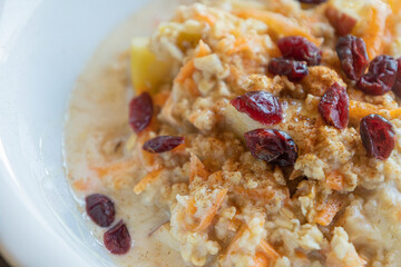  delicious and healthy oatmeal served in a white bowl. The oatmeal is topped with grated carrots, fresh apple slices, and dried red cranberries