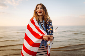 Young woman wrapped in American flag holding sparklers on sunset beach. Beautiful woman enjoying sunset. Concept of independence, celebration.