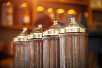 Clear glass dispensers with ornate gold tops stand in a warm, dimly lit shop, showcasing Turkish coffee. The focus is on the intricate details of these traditional containers.