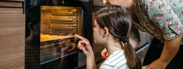 Banner of mother and daughter are carefully watching cookies baking in the oven, anticipating the delicious outcome of work. Young girl pointing with finger to biscuits. Family time cooking concept.