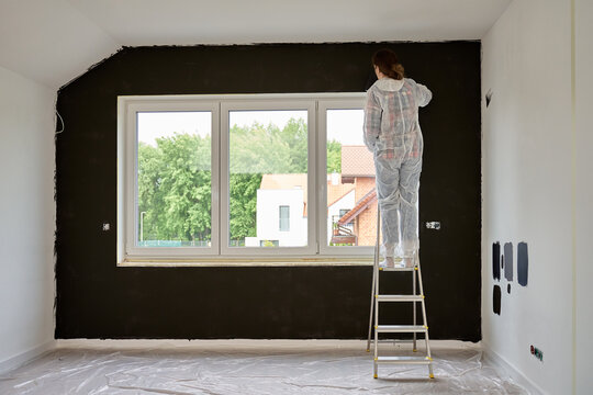 Woman in protective suit stands on ladder and paints wall black with roller in room under renovation. Interior painting process during home improvement