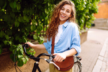 Cheerful woman with bicycle on sunny city street enjoying a walk. Tourist with bicycle walking in...