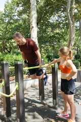 Man doing dips assisted by a woman at an outdoor gym.  They're exercising together in a park, enjoying a sunny day. Fitness, training, and teamwork are evident.