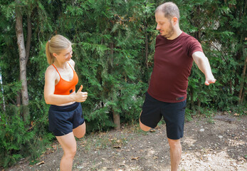 A woman gives a thumbs-up to a man during a leg-stretching exercise outdoors.  They are both wearing athletic wear.  Perfect for fitness, sports, or healthcare marketing.