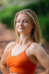 Portrait of a young blonde woman in an orange sports bra, smiling confidently.  She's outdoors with a blurred green background, suggesting a park or natural setting. The image evokes health, fitness,