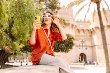Fototapeta premium Portrait of happy woman in hat taking selfie on sunny city old streets background. Beautiful tourist blogs on phone, enjoys sunset walk. Tourism, blogging concept.