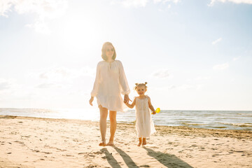 Happy mother and daughter strolling together on a sunny beach. Beautiful woman with her child enjoying the sunset on the beach overlooking the seascape. The concept of family, fun and childhood.