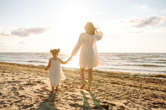 Happy mother and daughter strolling together on a sunny beach. Beautiful woman with her child enjoying the sunset on the beach overlooking the seascape. The concept of family, fun and childhood.