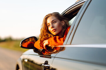 Cheerful woman leaning out of her car window feeling free. Young woman traveling by car enjoying...