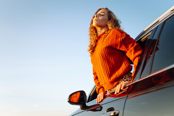 Cheerful woman leaning out of her car window feeling free. Young woman traveling by car enjoying...