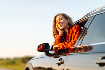 Naklejka premium Cheerful woman leaning out of her car window feeling free. Young woman traveling by car enjoying road adventure from car window. Adventure and transport concept.