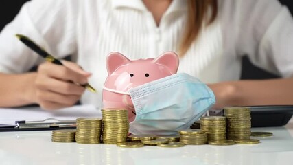 Close Up Pink Piggy Bank with Surgical Mask and Gold Coins with Businesswoman Writing on White Desk Finance Concept - Powered by Adobe