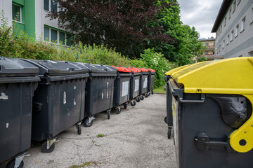 Multiple garbage containers with black, red, and yellow lids near residential buildings 