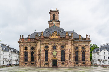 A large building Baroque church in the city of Saarbrucken in Germany with a clock tower and a small courtyard in front of it
