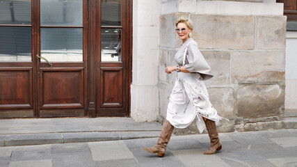 Fashionable mature woman walking city wearing asymmetrical gray loose-fit blouse, high-waisted white midi skirt with floral prints, suede knee-high boots, silver accessories and oversized sunglasses