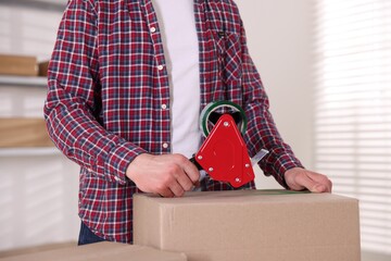 Man using dispenser with adhesive tape to seal cardboard box indoors, closeup