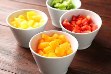 Pieces of fresh colorful bell peppers in bowls on wooden table, closeup