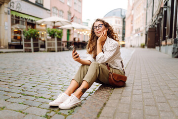 Fototapeta premium Portrait of a beautiful woman in glasses holding a phone on a sunset street. Beautiful woman texting on her phone, blogging and enjoying a walk in the city. Tourism concept.