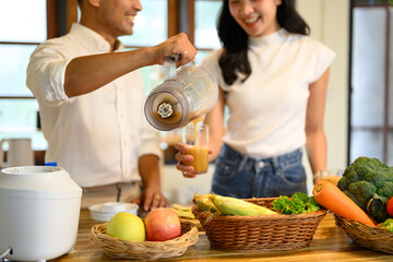 Smiling couple pouring fresh blended smoothie into a glass in a cozy kitchen