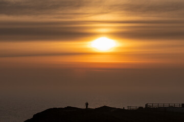 A solitary photographer stands silhouetted on a clifftop, gazing at the magnificent sunrise over...