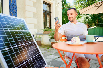 Young man charging smartphone from solar panel while sitting in cafe. Handsome male drinking coffee and checking his phone next to solar battery. Conscious male choosing sustainable lifestyle.
