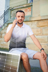 Handsome man using smartphone connected to photovoltaic solar panel. Integration of sustainable renewable energy into everyday life, demonstrating practical use of solar power for charging devices.