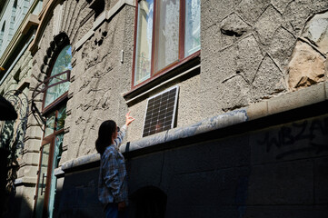 Woman holding photovoltaic solar panel in front of historical building, showing Ok sign. Concept of integration of sustainable renewable energy sources into old architecture.