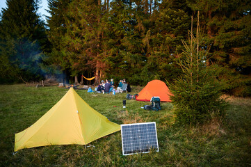 Solar panel in grassy field with tents in background highlights eco-friendly camping setup. Scene includes campers, emphasizing integration of renewable energy in outdoor activities.