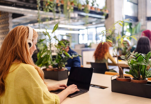 Young office worker wearing headphones typing on laptop in green workspace