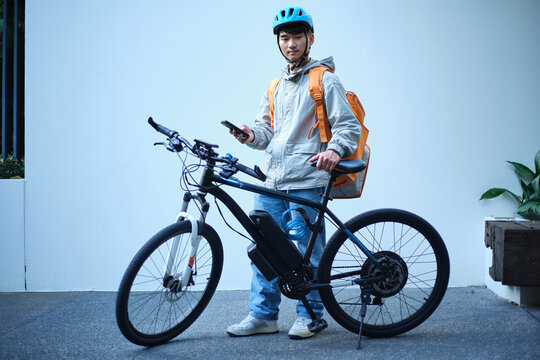 Young Asian delivery guy standing beside his bicycle holding a mobile phone
