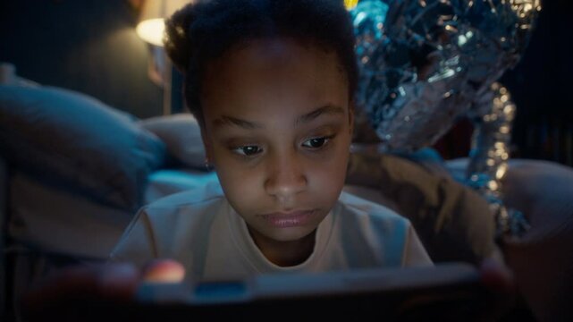Closeup shot of little African American girl sitting in dark room at night absorbed into smartphone screen while mysterious alien monster sneaking up behind her but remaining unnoticed