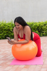 Latin woman using smartphone while doing pilates with an exercise ball outdoors