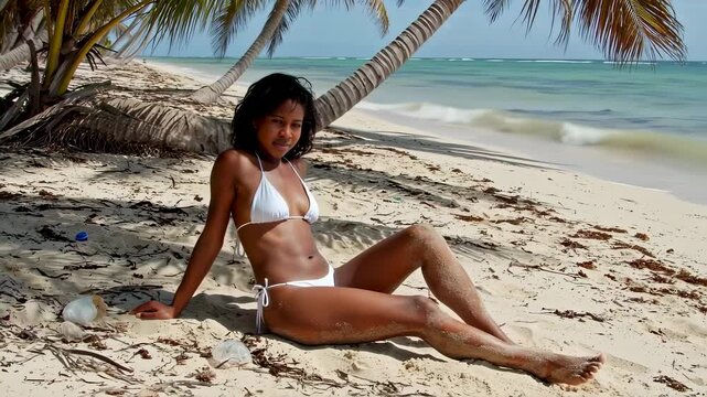 A beautiful young Black woman wearing a white string bikini sits gracefully on a sandy tropical shore.