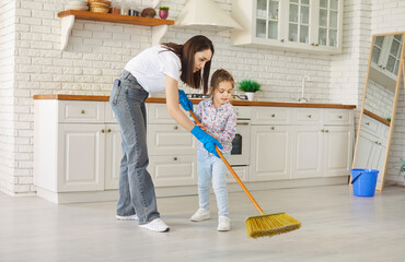 Loving mother and little helper daughter learning sweeping the floor together with broom wearing...